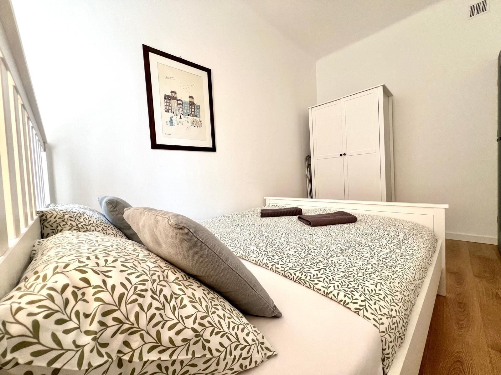 A bedroom with a bed, pillows, a wardrobe, and a framed picture on the wall.