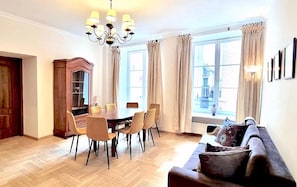 A dining area with a wooden table and chairs, a chandelier, and large windows with curtains. - AAA STAY Castle Square Old Town (Warsaw)