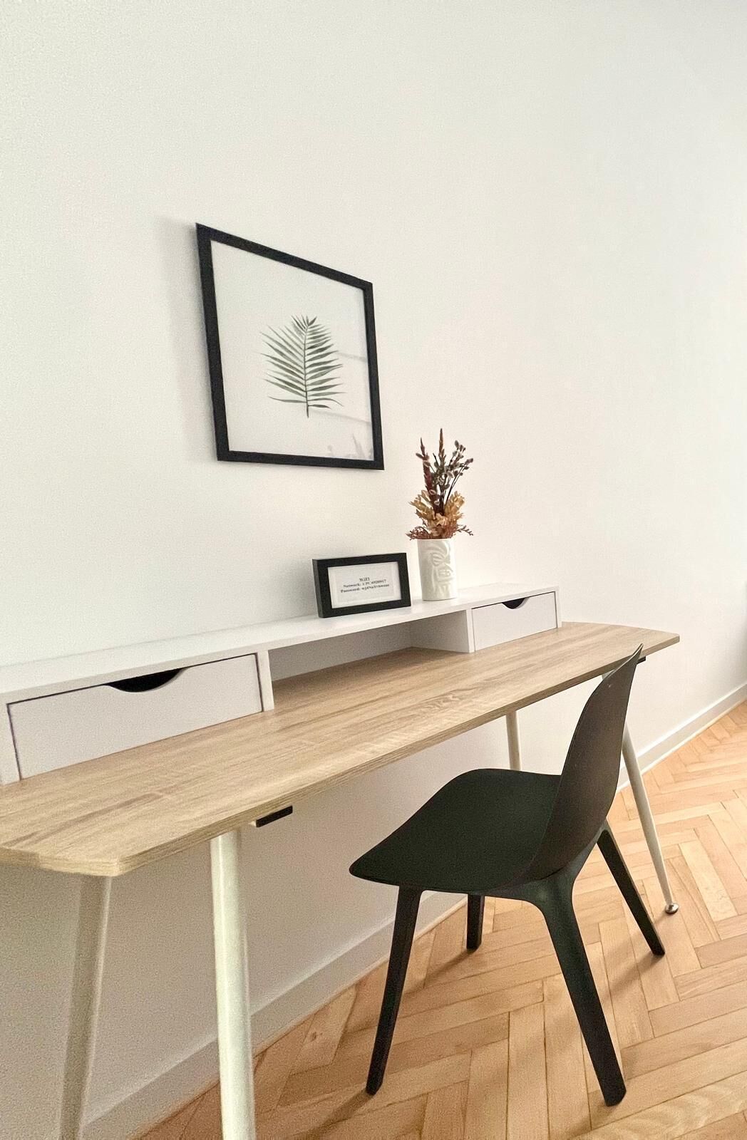 A modern desk with a wooden top and metal legs, a black chair, a white console unit with drawers, and a framed plant on the wall.