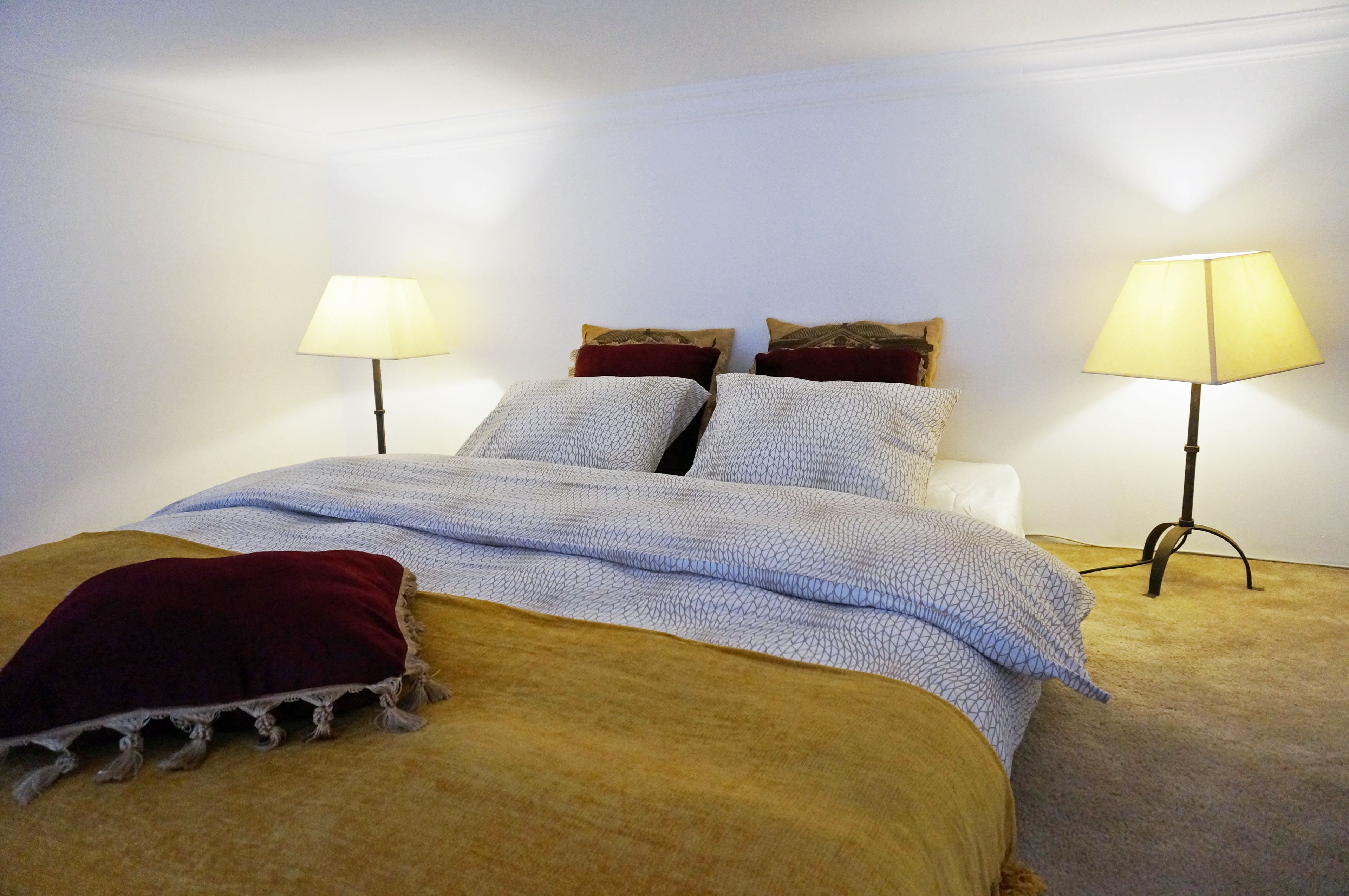 A neatly made bed with a patterned blanket, two pillows, and a red throw. There are two bedside lamps on either side of the bed.