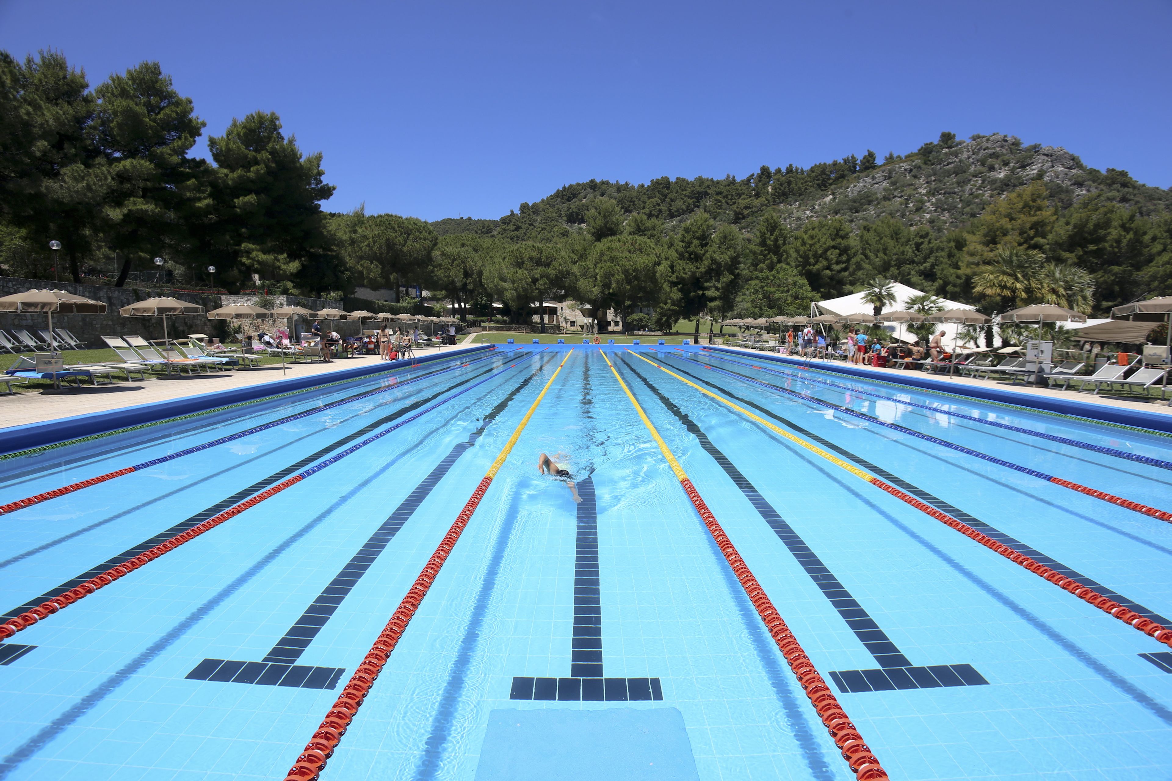 A swimming pool with multiple lanes, surrounded by lounge chairs and umbrellas, with a mountainous backdrop.