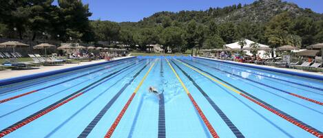 A swimming pool with multiple lanes, surrounded by lounge chairs and umbrellas, with a mountainous backdrop.