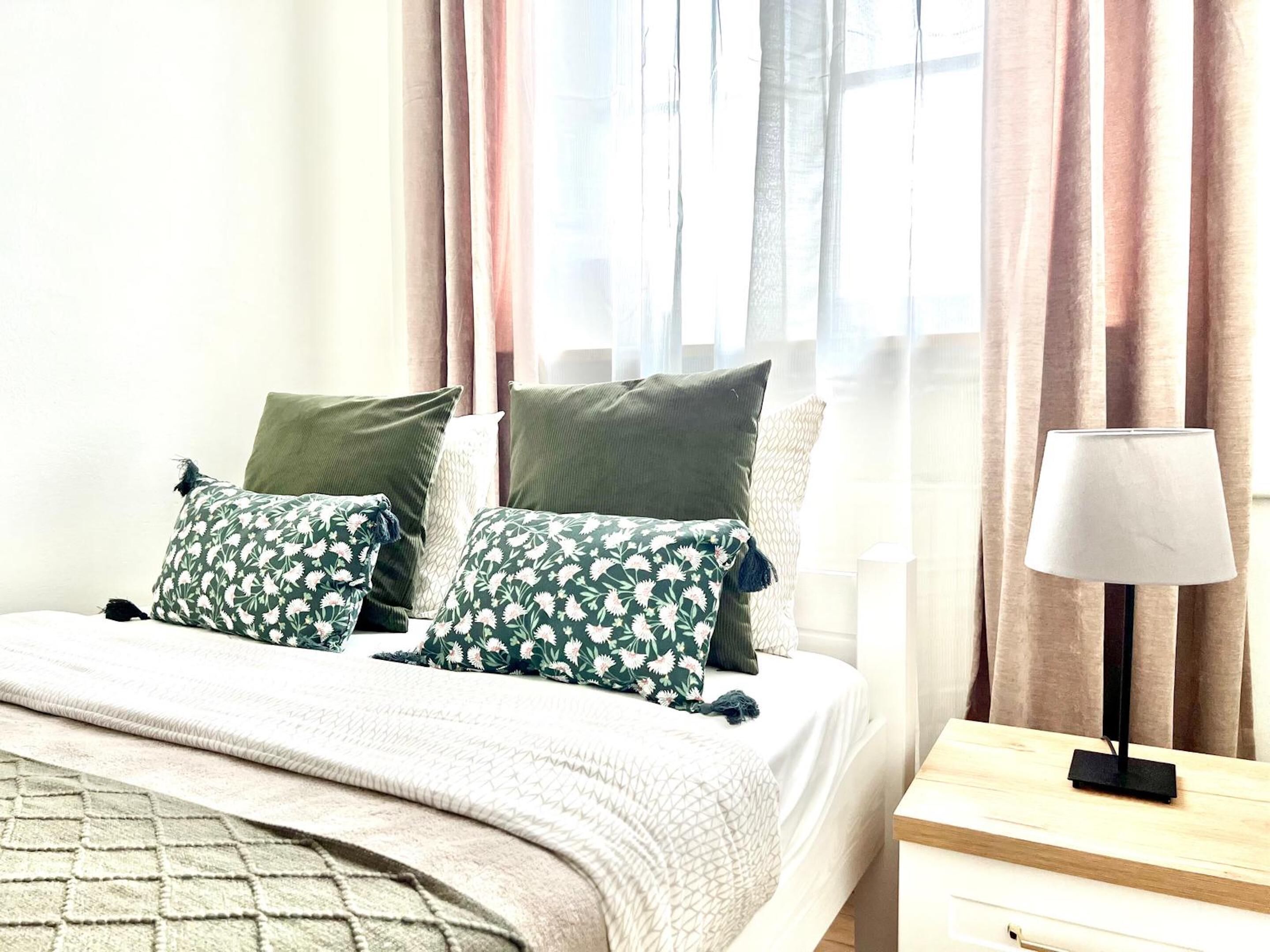 A neatly made bed with floral-patterned pillows, a bedside table with a lamp, and sheer curtains allowing natural light.