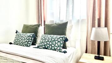 A neatly made bed with floral-patterned pillows, a bedside table with a lamp, and sheer curtains allowing natural light.