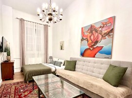 Una sala moderna con un sofá, una mesa de centro de vidrio, una cama con manta verde, una araña, un cuadro y una ventana con cortinas translúcidas.