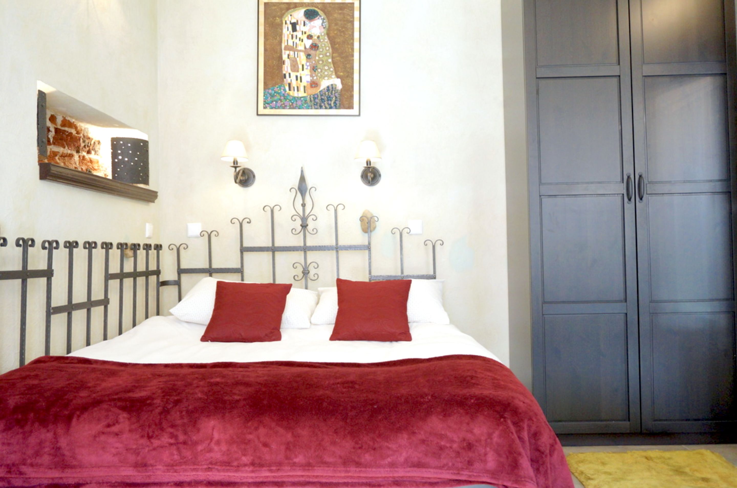 A bedroom with a bed, a red blanket, a painting, a wardrobe, and a small shelf with a picture.