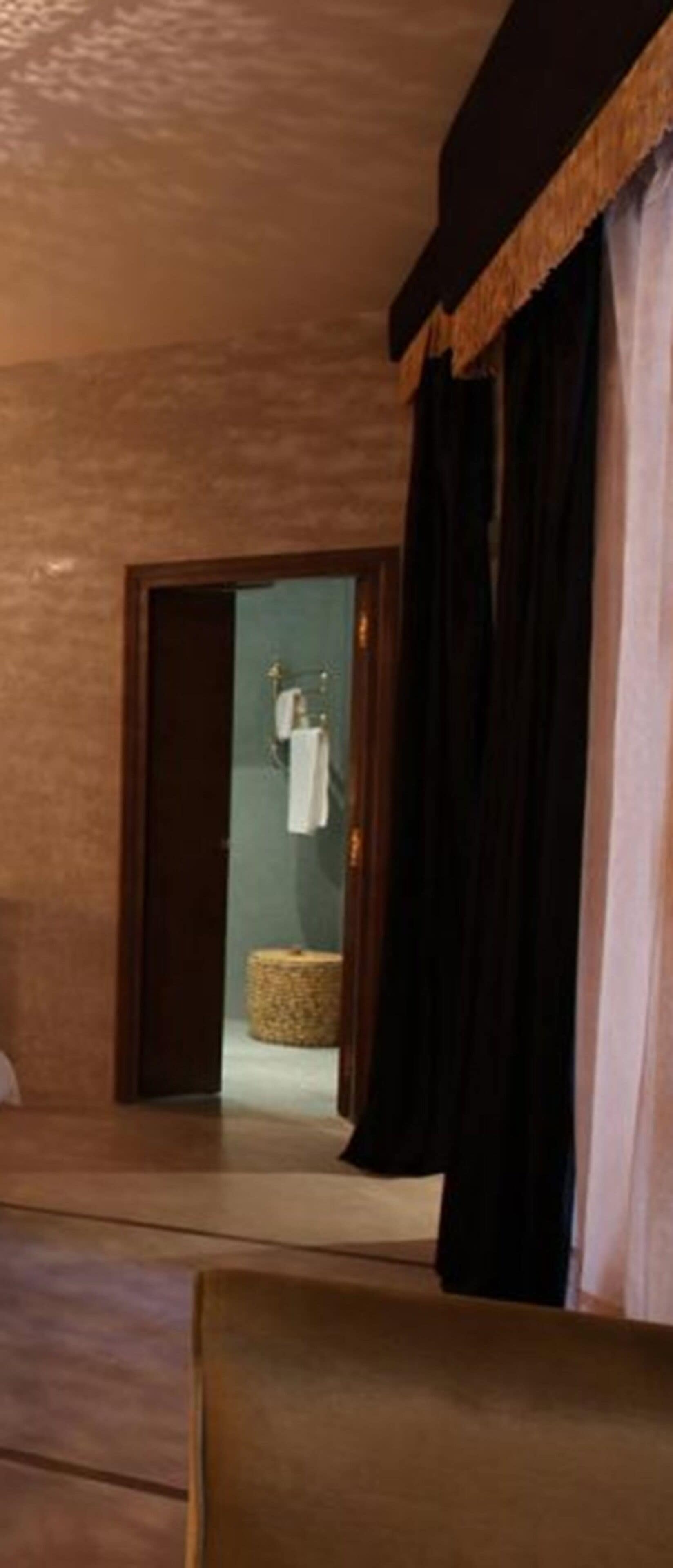 A hotel room with a wooden floor, a bed, a small round wicker basket, and a bathroom visible through an open door.