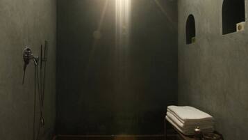 Una habitación estrecha y tenue, con zona de ducha, una mesita y una pila de toallas, y un lavamanos en la pared.