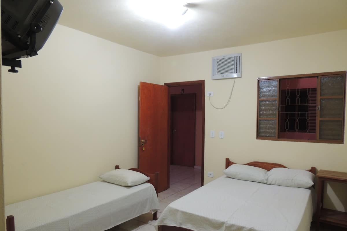 A room with two single beds, a wooden nightstand, a wall-mounted air conditioner, and a window with bars.
