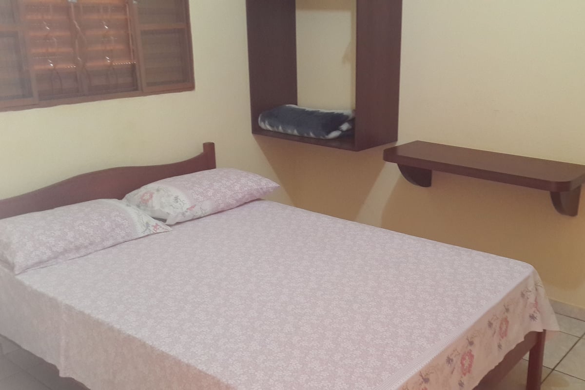 A bedroom with a bed, a nightstand, a wall-mounted shelf, and a window with blinds.