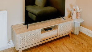 A television on a wooden cabinet with a remote control drawer, a white macrame wall hanging, and a small white table with decorative items. - Inverness Holiday House (Inverness)