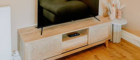 A television on a wooden cabinet with a remote control drawer, a white macrame wall hanging, and a small white table with decorative items.