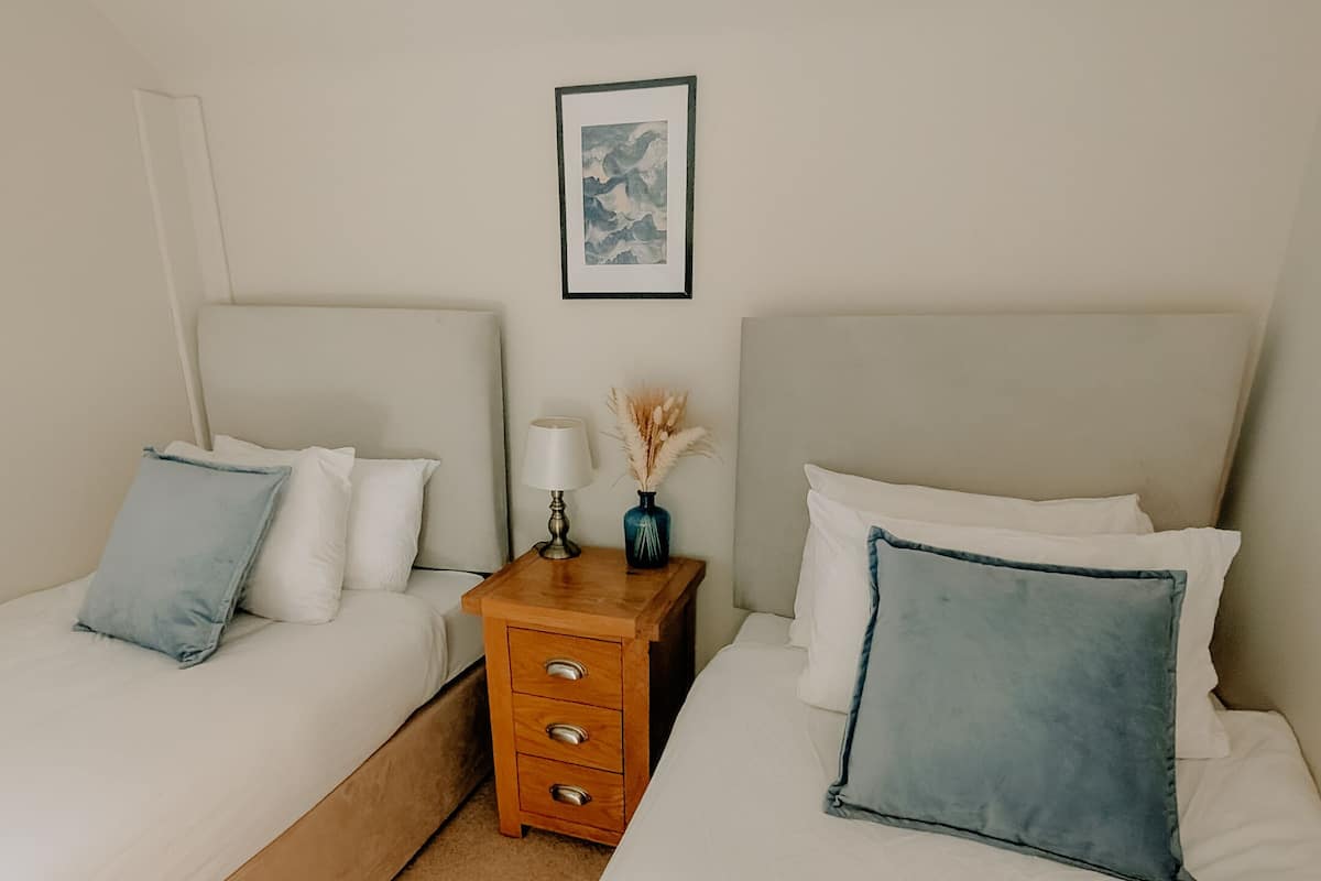 Une chambre double avec deux lits, une table de chevet, une lampe, un vase avec des fleurs séchées et un tableau encadré au mur.