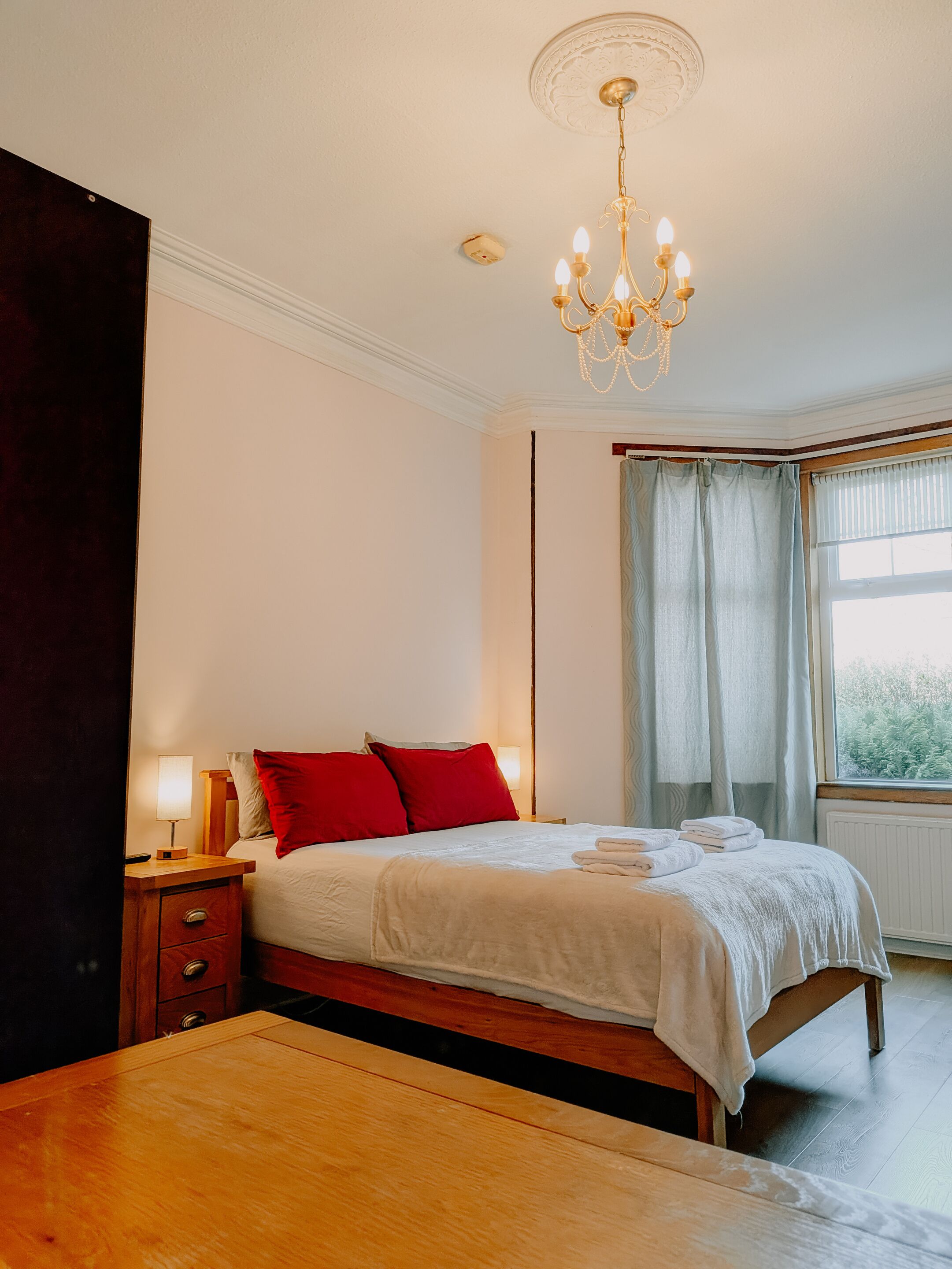 Une chambre à coucher avec un lit, des tables de chevet, un banc en bois, un lustre, une fenêtre avec des rideaux et une vue sur l’extérieur.
