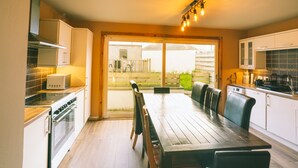 A modern kitchen with a wooden dining table, white cabinets, and a view through the glass door to the outside. - Inverness Holiday House (Inverness)