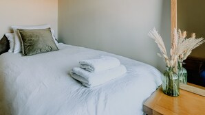 A neatly made bed with white linens and a folded towel, a wooden bedside table with a vase of dried flowers, and a large mirror on the wall. - Inverness Holiday House (Inverness)