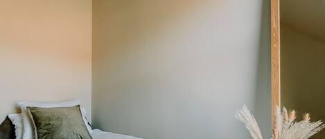 A neatly made bed with white linens and a folded towel, a wooden bedside table with a vase of dried flowers, and a large mirror on the wall.
