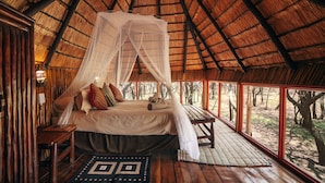 Ein Schlafzimmer mit Strohdach, einem Baldachinbett, Holzmöbeln und Blick auf den Wald.
