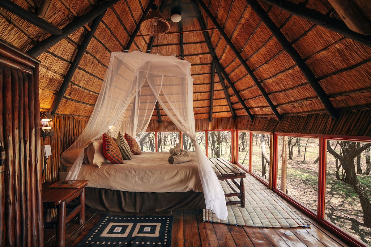 A thatched-roof bedroom with a canopy bed, wooden furniture, and a view of the forest.