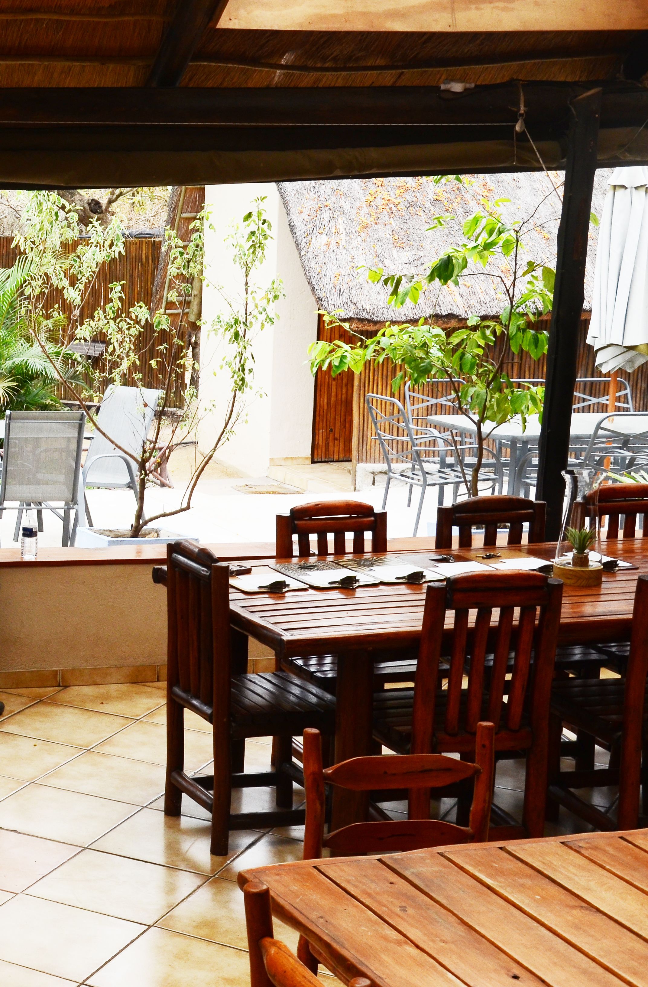 An outdoor dining area with wooden tables and chairs, a thatched roof, and potted plants.