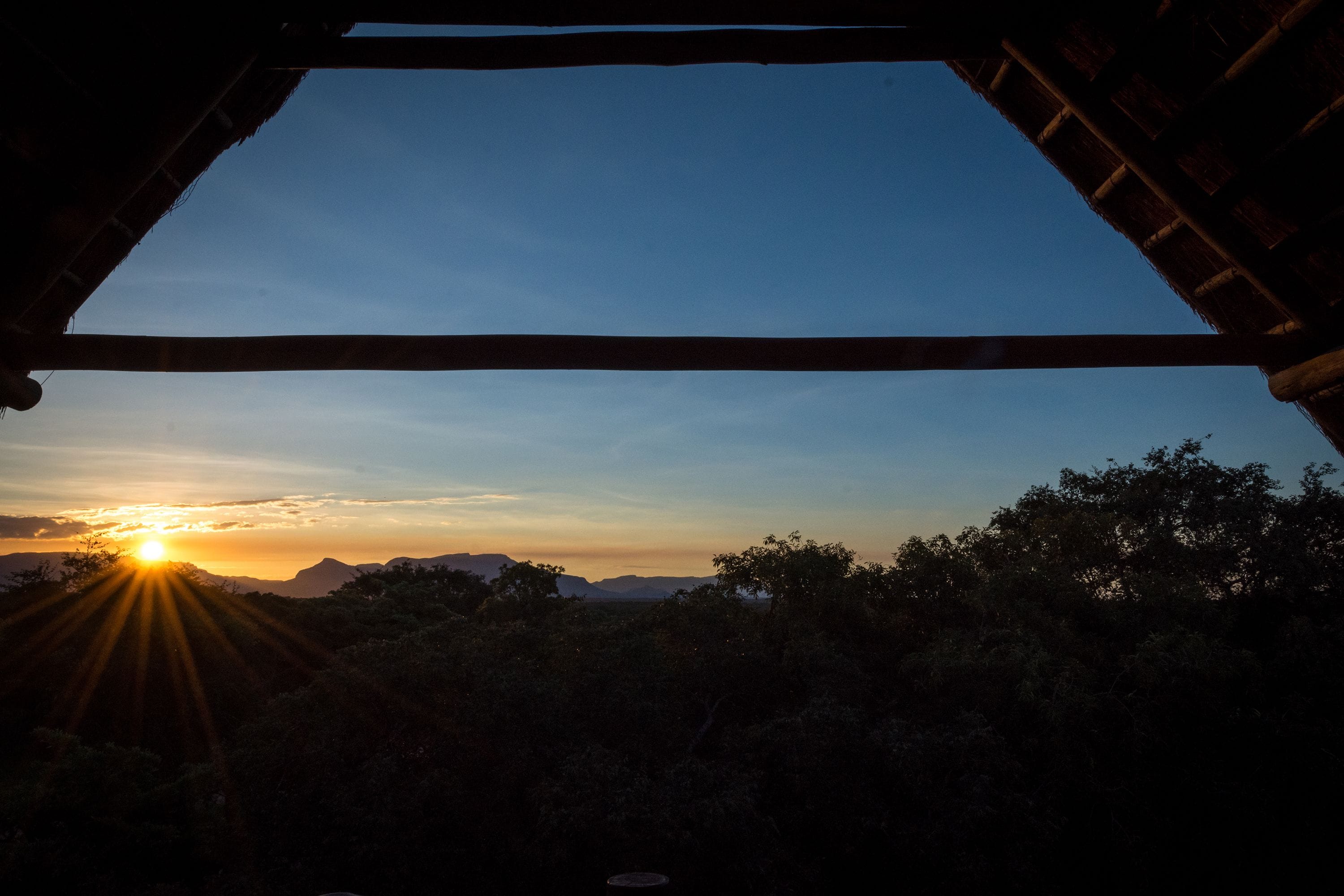 Une vue par une fenêtre au coucher du soleil, avec le soleil se cachant derrière une chaîne de collines et d’arbres.