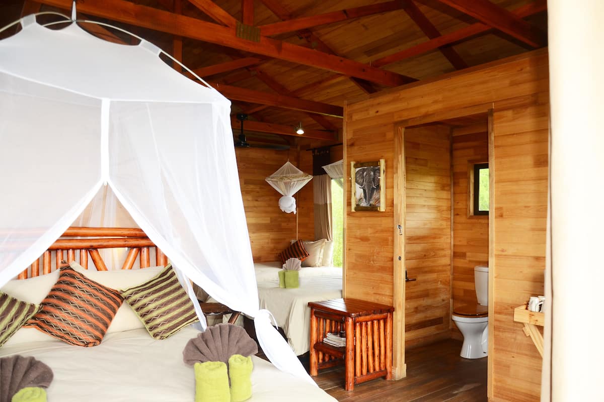 A wooden cabin with a bed under a mosquito net, a small table, and a toilet.