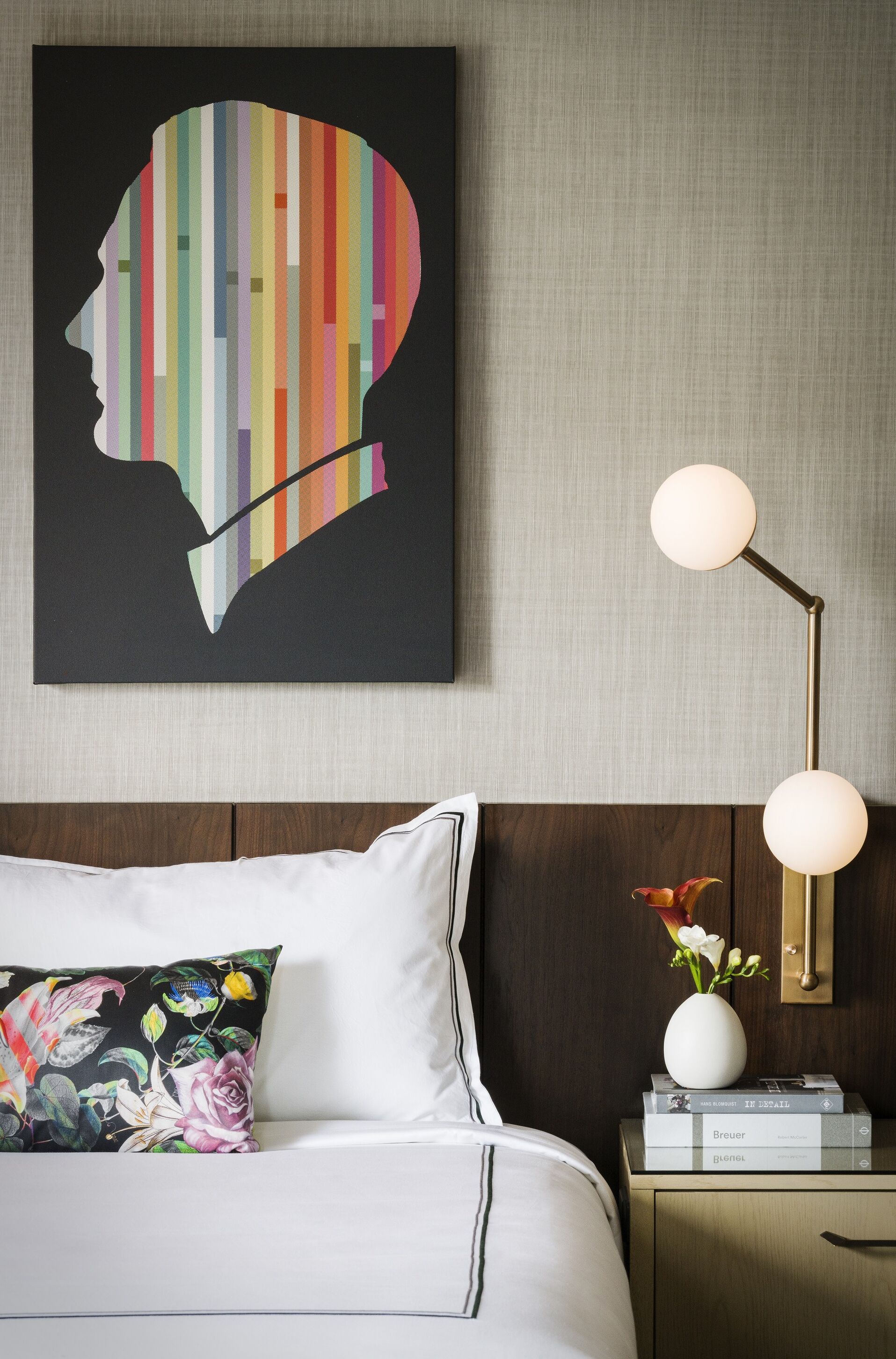 Un lit bien fait, avec du linge de lit blanc, un coussin à motifs floraux et une tête de lit moderne. Sur le mur, on peut voir une œuvre d’art représentant le profil d’une personne, composé de rayures verticales colorées. À côté du lit, il y a une table de chevet avec une lampe et un vase rempli de fleurs.