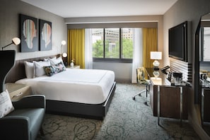 A hotel room with a large bed, a desk with a lamp, a TV, and artwork on the wall.