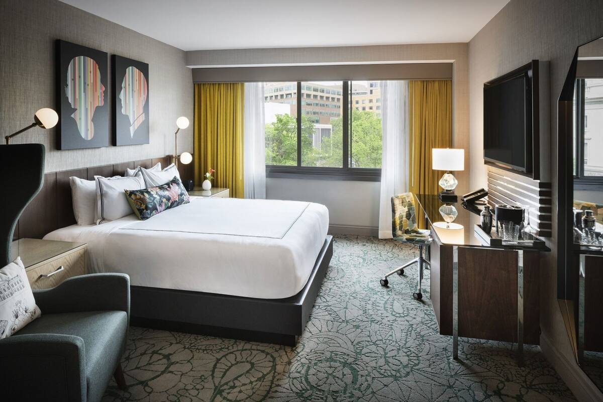 A hotel room with a large bed, a desk with a lamp, a chair, and artwork on the wall.