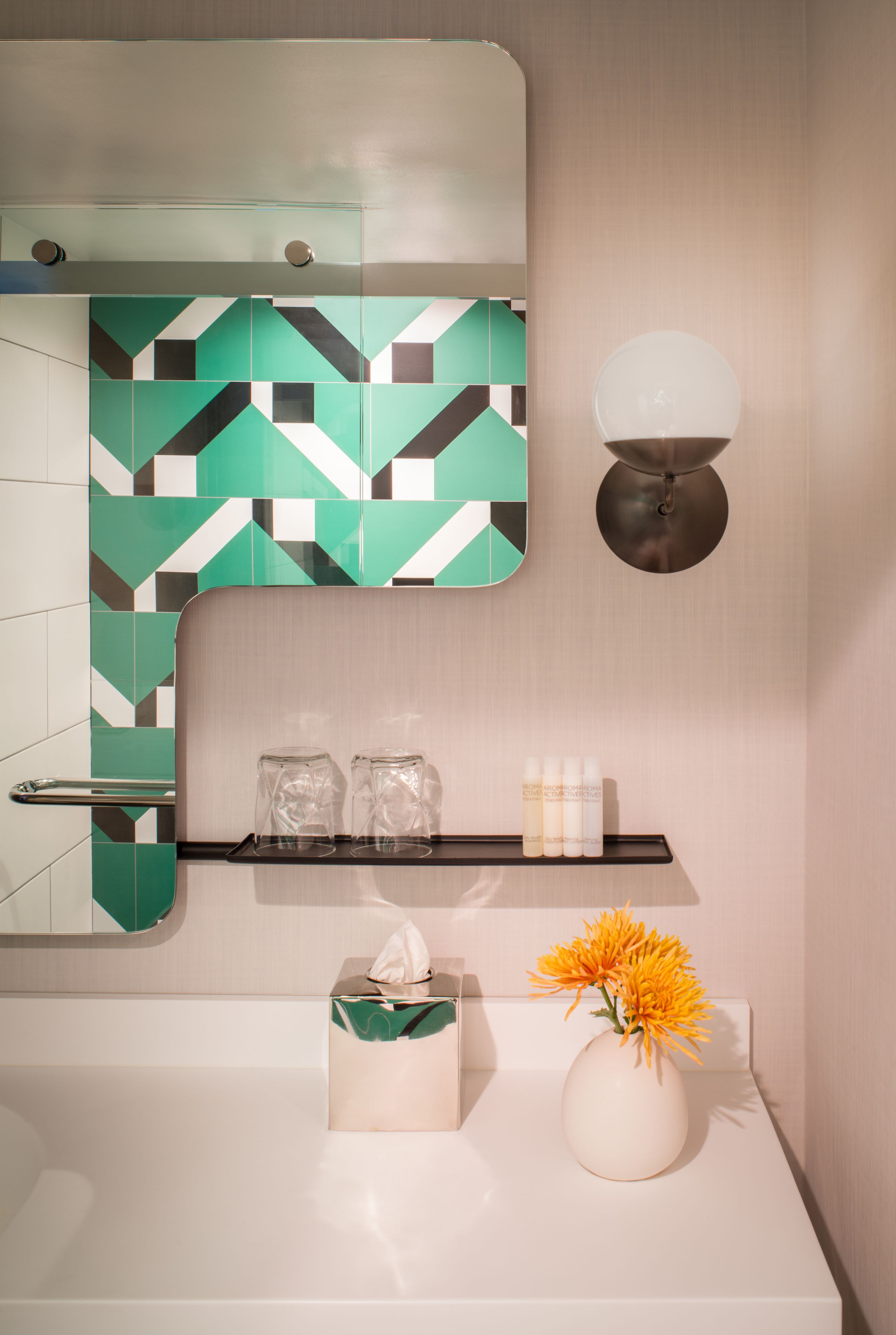 A bathroom with a geometric tile backsplash, a mirror, a wall-mounted light fixture, a shelf with toiletries, a tissue box, and a vase with flowers.