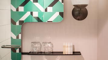 A bathroom with a geometric tile backsplash, a mirror, a wall-mounted light fixture, a shelf with toiletries, a tissue box, and a vase with flowers.