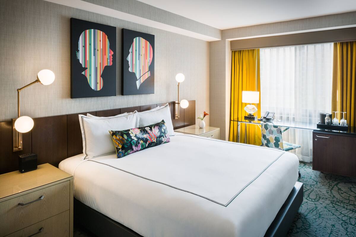 A hotel room with a large bed, two bedside tables, a nightstand, a desk with a lamp, a chair, a window with curtains, and artwork on the wall.