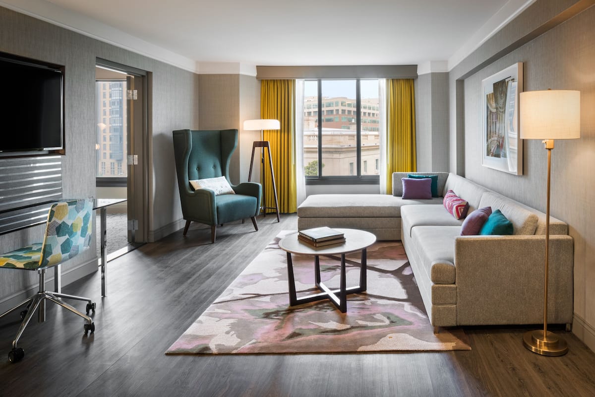 A hotel room with a bed, a sofa, a desk with a chair, a standing lamp, and a TV.