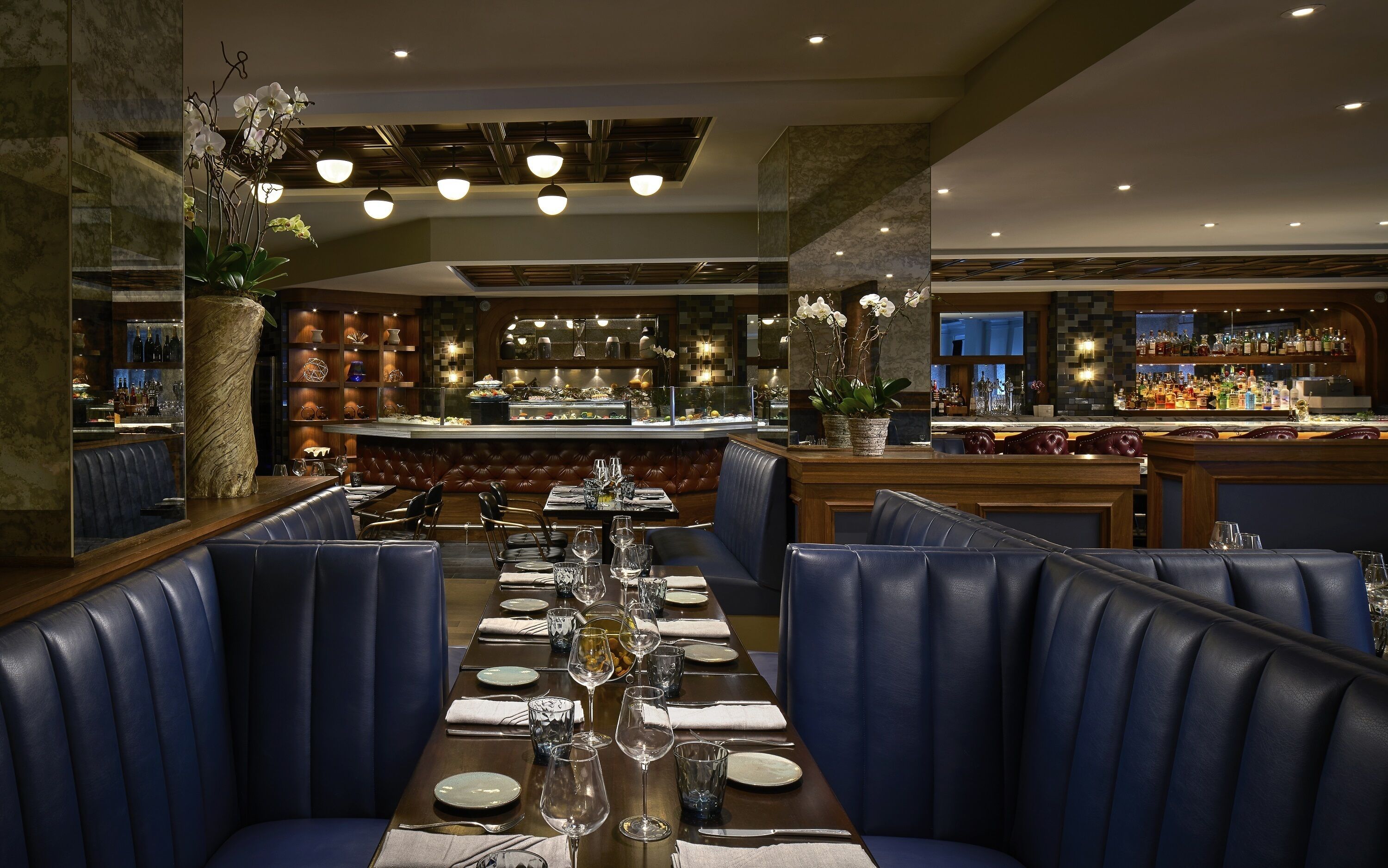 A restaurant with a long dining table set for a meal, surrounded by leather booths, and a bar area with shelves of bottles.