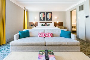 Une chambre d’hôtel avec un canapé, un lit, deux œuvres d’art murales, une table de chevet avec une lampe et une télécommande posée sur un magazine.
