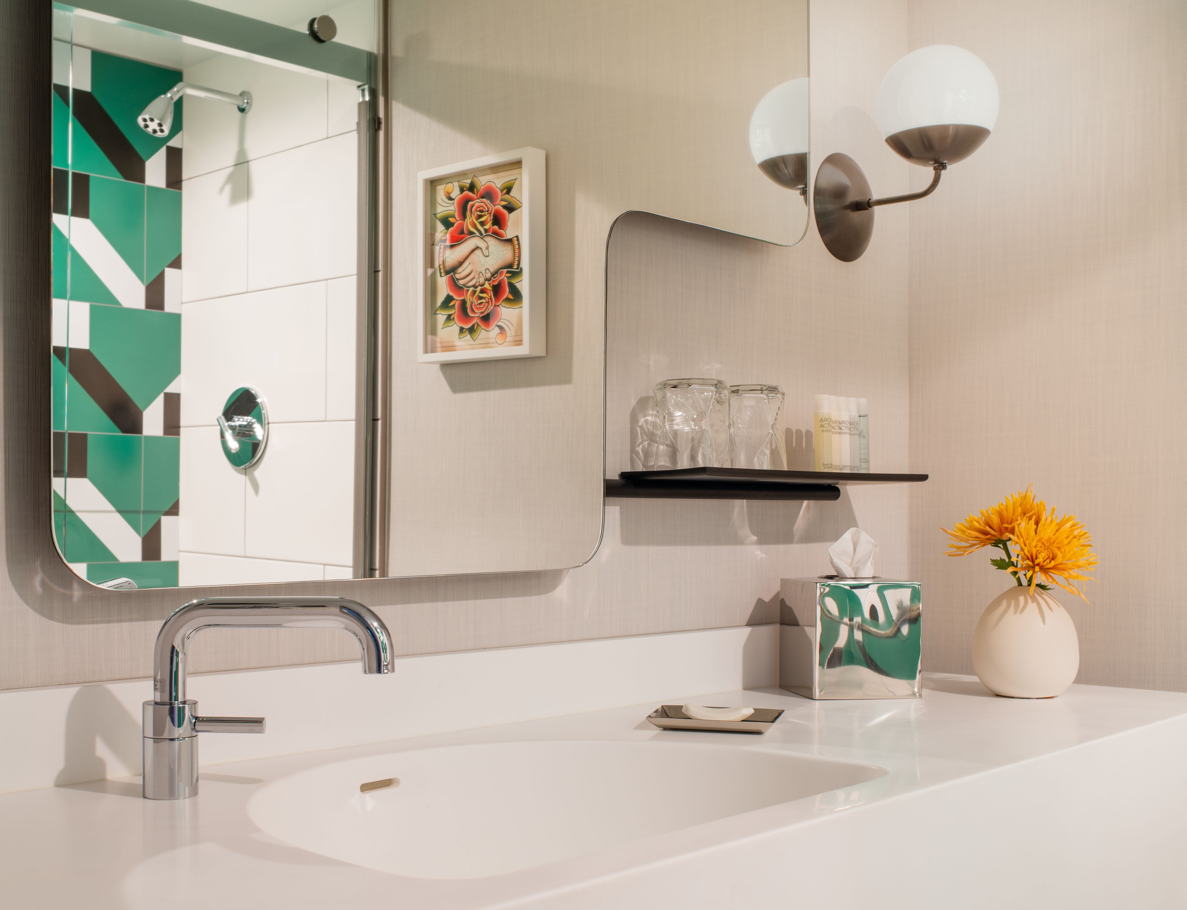 A bathroom with a modern sink, a mirror, a towel dispenser, a shelf with toiletries, and a vase with flowers.