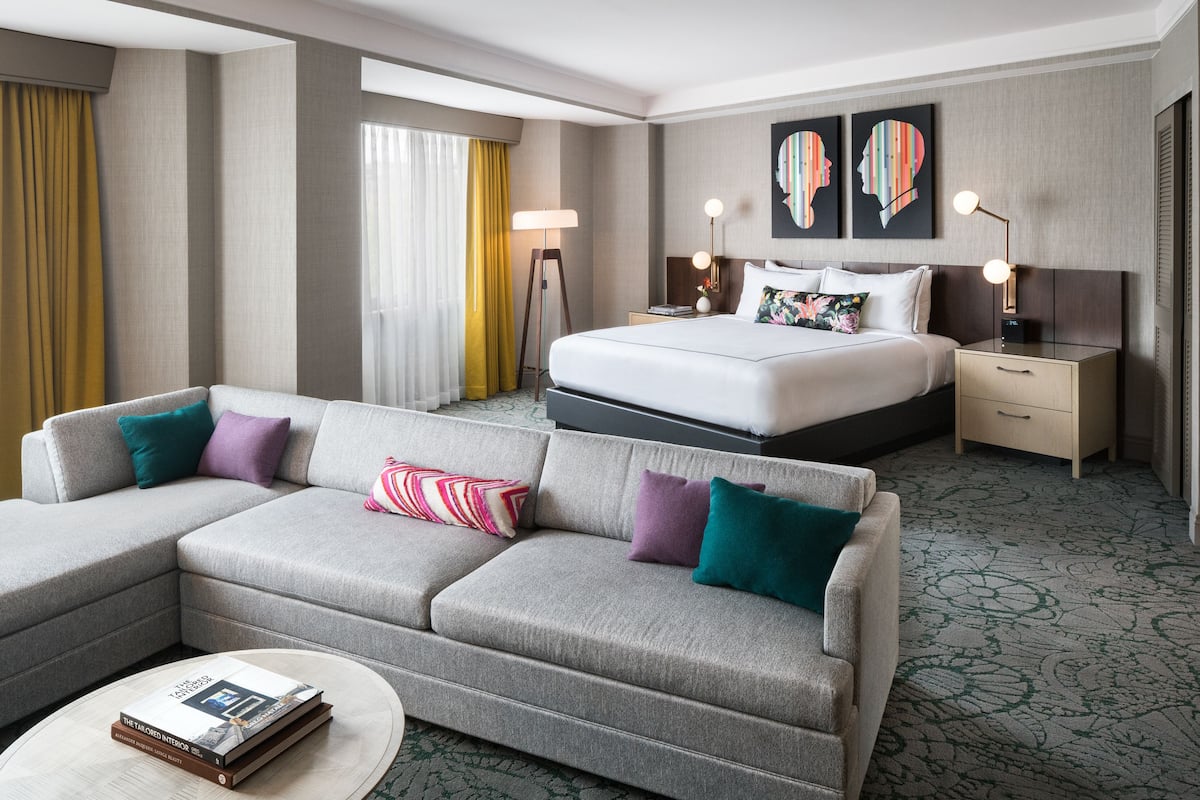 A hotel room with a large bed, a grey sofa, a bedside table, and a nightstand.