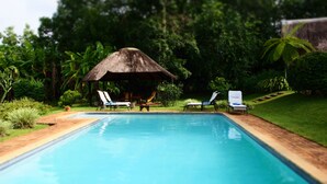 Una piscina con sillas de descanso y una cabaña con techo de paja en un exuberante jardín.