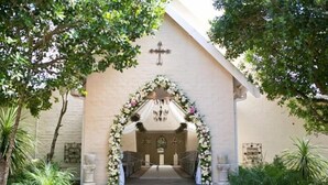 L’entrée d’une église, surmontée d’une croix au-dessus de la porte, avec une arche ornée de fleurs et des marches menant à l’entrée.