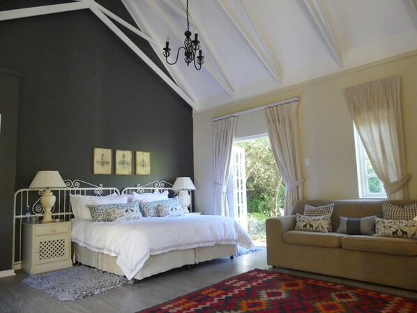 A bedroom with a bed, bedside tables, a sofa, a chandelier, and a window with curtains. - The Plantation (Gqeberha (Port Elizabeth))
