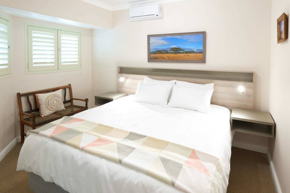 A neatly arranged bedroom with a large bed, a nightstand, a wooden chair, and a wall-mounted air conditioner.