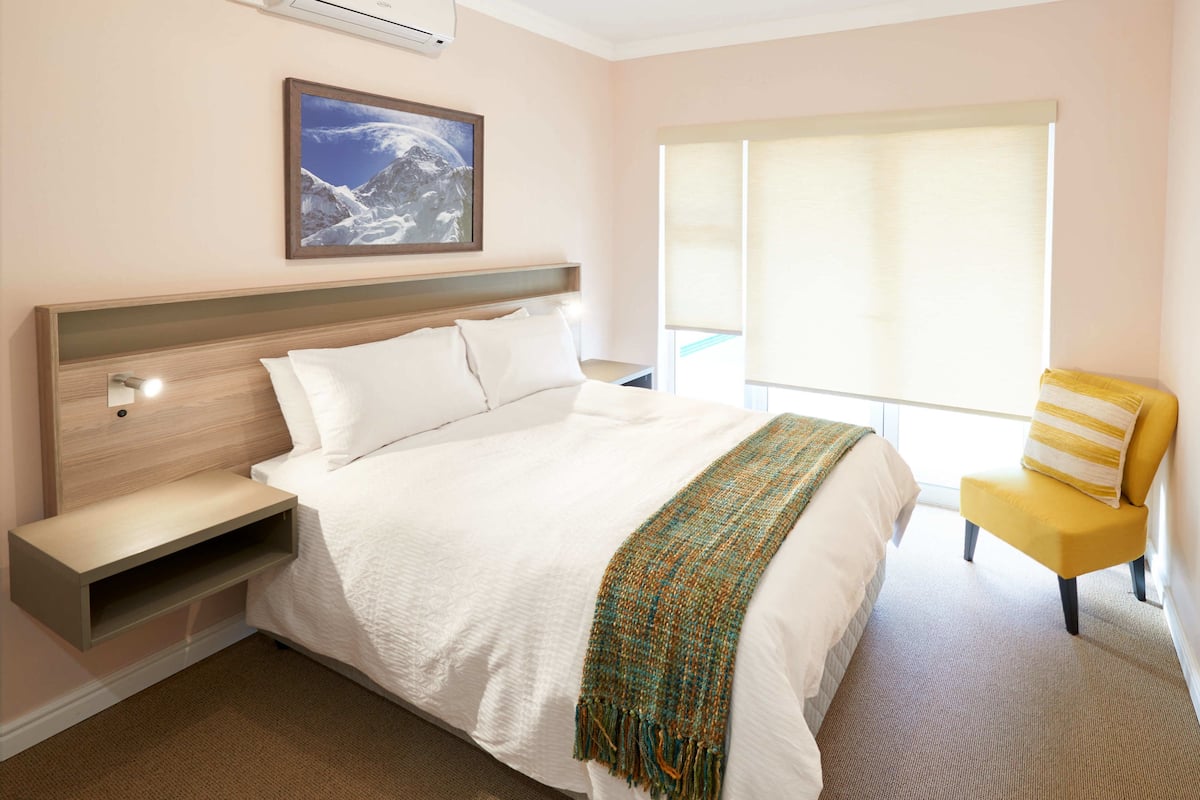 A hotel room with a bed, a nightstand, a chair, a window with blinds, and a painting on the wall.