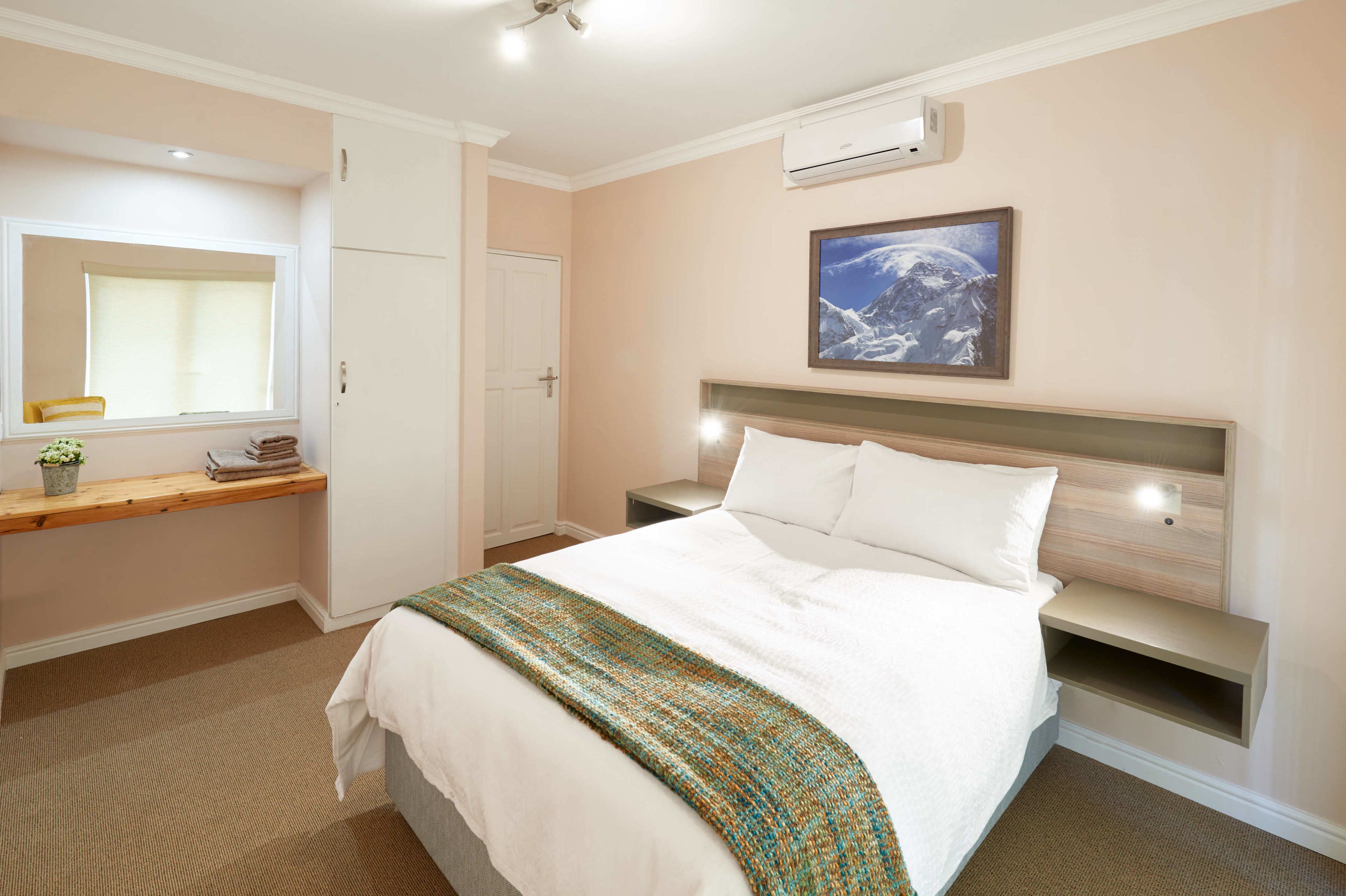 A hotel room with a bed, a bedside table, a nightstand, a window with a view, and an air conditioning unit.
