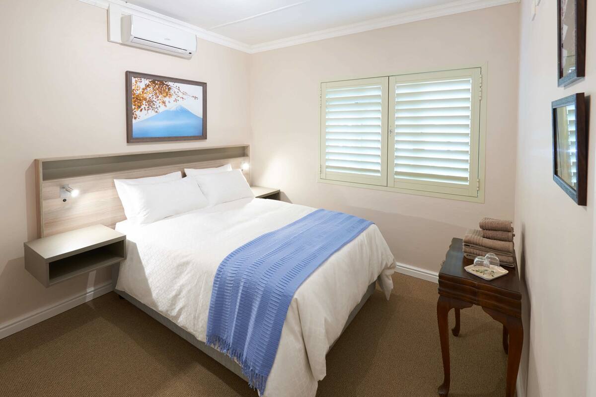 A neatly arranged hotel room with a bed, bedside tables, a nightstand, a window with blinds, and a wall-mounted air conditioner.