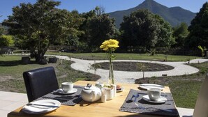 A dining table set with plates, cups, and a vase of yellow flowers, overlooking a landscaped garden and a mountain. - Tsitsikamma Manor (Storms River)