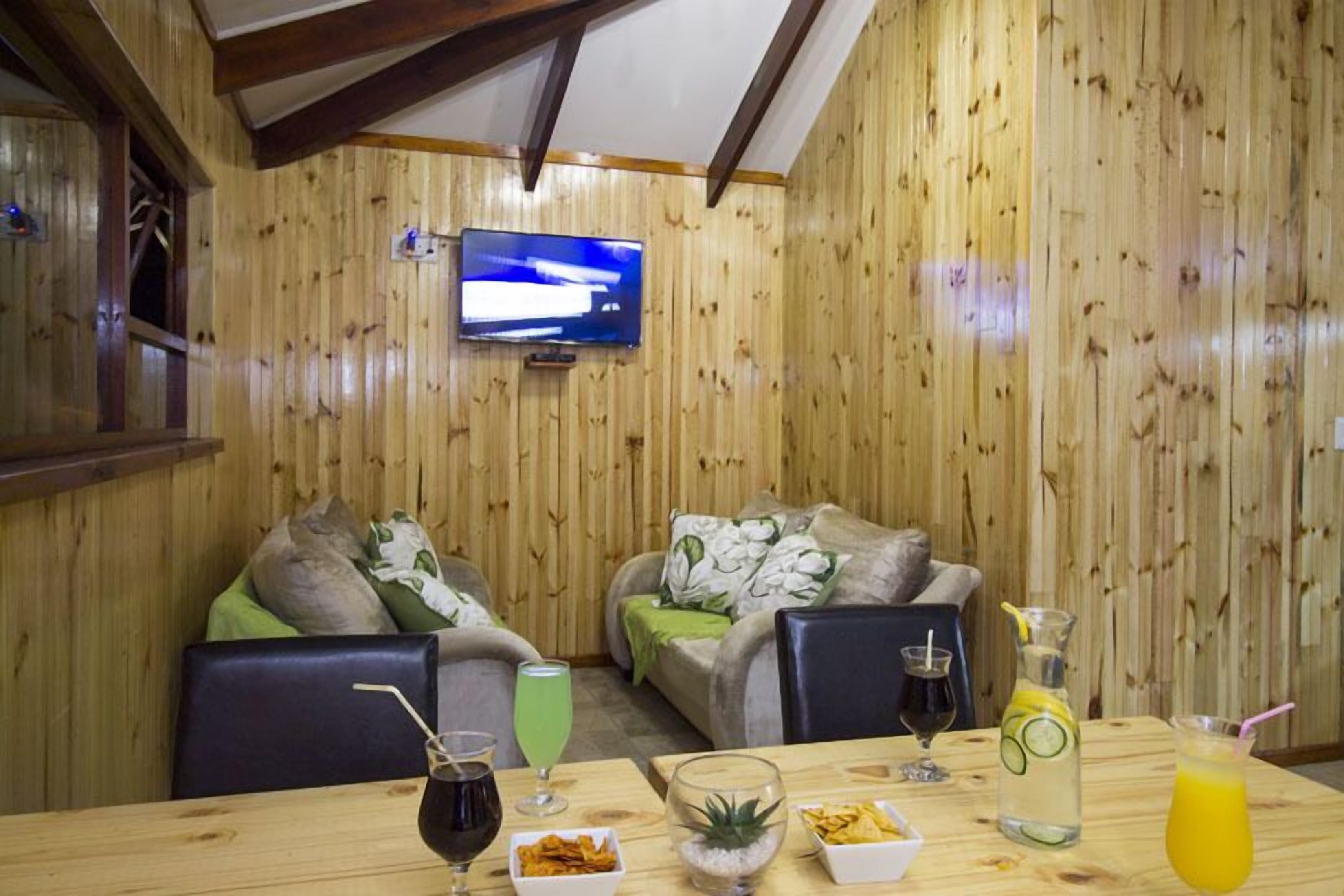 Una habitación acogedora con paredes de madera, un televisor de pantalla plana montado en la pared, dos sofás con cojines, una mesa de comedor con bebidas y aperitivos, y una ventana con persianas.