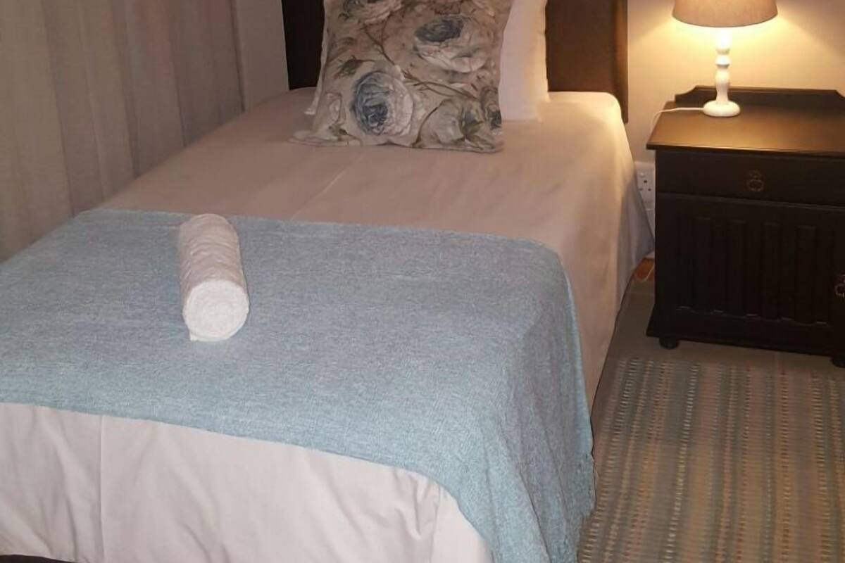 A neatly made bed with a blue blanket and a rolled towel, a dark headboard, a floral pillow, and a bedside lamp on a wooden nightstand.