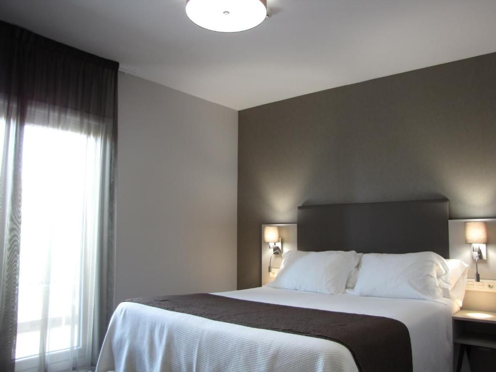 Habitación de hotel con una cama grande, dos lámparas de noche, una ventana con cortinas e iluminación en el techo.
