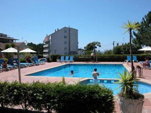 Ein Schwimmbecken mit Liegestühlen und Sonnenschirmen, umgeben von viel Grün und einem Gebäude im Hintergrund.
