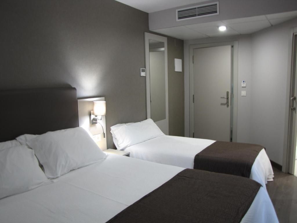 Una habitación de hotel con dos camas, una mesita de noche con lámpara, un espejo y una puerta.